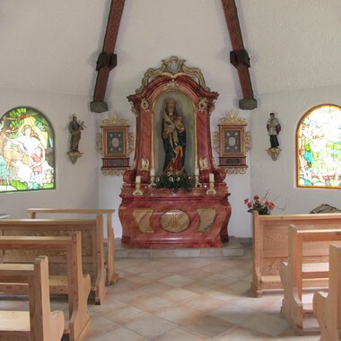 A small chapel with wooden bench rows and colorful windows. In the foreground stands an ornate altar with a statue. | © Stefan Heim