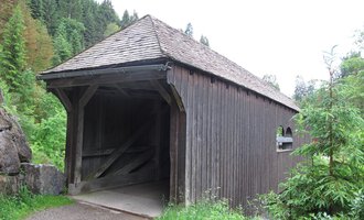An old wooden bridge in the midst of a green forest area. The bridge has a pointed roof and is surrounded by trees. | © Stefan Heim
