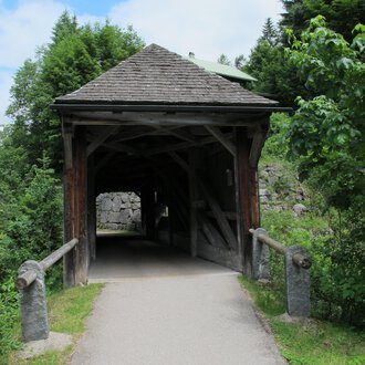 Eine Holzbrücke führt durch einen überdachten Gang. Umgeben von grünen Bäumen und einem klaren Himmel wirkt die Umgebung friedlich. | © Stefan Heim