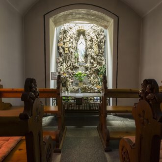 Eine stille Kapelle mit Holzbänken und einer Statue in der Nische. Die Wände sind sanft beleuchtet und schaffen eine friedliche Atmosphäre. | © Kleinwalsertal Tourismus | Oliver Farys