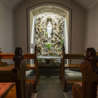 Eine stille Kapelle mit Holzbänken und einer Statue in der Nische. Die Wände sind sanft beleuchtet und schaffen eine friedliche Atmosphäre. | © Kleinwalsertal Tourismus | Oliver Farys