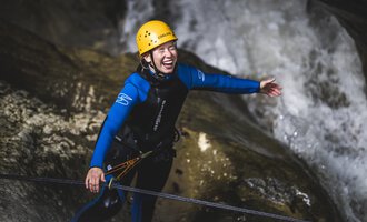 Eine Person in einem blauen Neoprenanzug und gelben Helm steht in einer felsigen Umgebung. Sie lächelt und sieht glücklich aus, während Wasser im Hintergrund fließt. | © MAP-Erlebnis