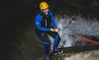 Eine Person in einem blauen Neoprenanzug und einem Helm steht an einem Wasserfall. Sie zieht mit einem Seil und hat ein breites Lächeln im Gesicht. | © MAP-Erlebnis