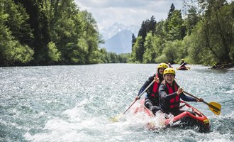 Eine Gruppe von Personen paddelt mit einem Raft auf einem klaren Fluss. Im Hintergrund sind Bäume und Berge sichtbar. | © MAP-Erlebnis