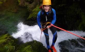 Ein abenteuerlicher Mensch beim Abseilen an einem kleinen Wasserfall. Er trägt einen Helm und einen Neoprenanzug und lächelt in die Kamera. | © MAP-Erlebnis