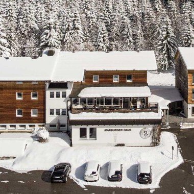 Eine schneebedeckte Landschaft mit mehreren Gebäuden im Chalet-Stil. Umgeben von hohen, schneebedeckten Tannenbäumen. | © Marburger Haus | Sweetchili