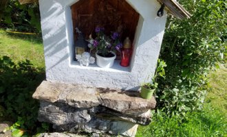 Eine kleine weiße Kapelle mit einem Giebeldach steht auf einem Steinfundament. Umgeben von grünem Gras und Bäumen ist sie mit Blumen dekoriert. | © Maxhütte | Viera Krafcik