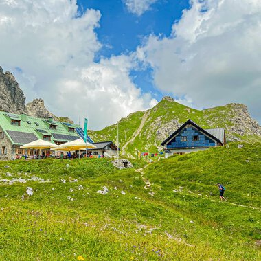 Eine malerische Berglandschaft mit gemütlichen Hütten und grünen Wiesen. Im Hintergrund sind beeindruckende Felsen und ein blauer Himmel mit Wolken zu sehen. | © Mindelheimer Hütte | Wolfgang Stache