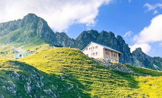 Ein typisches Berghaus steht auf einer grünen Wiese mit majestätischen Bergen im Hintergrund. Der Himmel ist blau und teilweise bewölkt. | © Mindelheimer Hütte | Wolfgang Stache