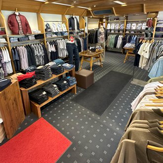 Ein modischer Bekleidungsladen mit einer Vielzahl von Hemden und Pullovern. Die Regale sind ordentlich sortiert und der Boden ist mit einem roten Teppich sowie einem gemusterten Teppich ausgelegt. | © Moosbrugger Mode e.U.