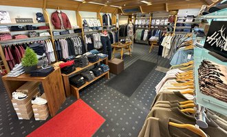 Ein modischer Bekleidungsladen mit einer Vielzahl von Hemden und Pullovern. Die Regale sind ordentlich sortiert und der Boden ist mit einem roten Teppich sowie einem gemusterten Teppich ausgelegt. | © Moosbrugger Mode e.U.