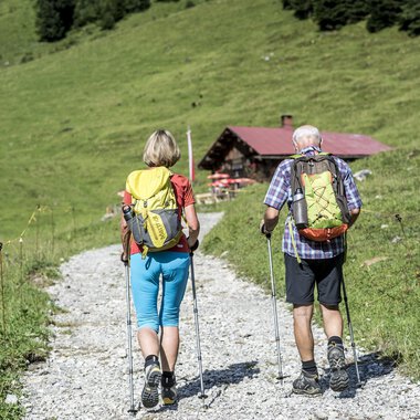 Ein Paar wandert auf einem Kiesweg in einer grünen Landschaft. Im Hintergrund ist eine kleine Hütte zu sehen. | © Kleinwalsertal Tourismus | Dominik Berchtold