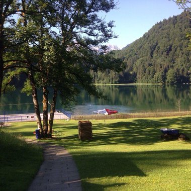Ein ruhiger See umgeben von Bäumen und grünen Wiesen. Im Hintergrund sind Berge und Sonnenliegen am Ufer zu sehen. | © Kleinwalsertal Tourismus | Naturbad Freibergsee
