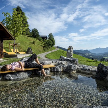 Eine entspannende Szene in den Bergen mit einer Person, die am Ufer eines kleinen Teiches liegt. Im Hintergrund sind grüne Hügel und ein klarer Himmel zu sehen. | © Kleinwalsertal Tourismus | Dominik Berchtold