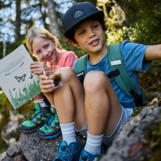 Zwei Kinder sitzen auf einem Stein in der Natur. Eines der Kinder hält ein Buch, während das andere ein Glas mit etwas in der Hand hat.