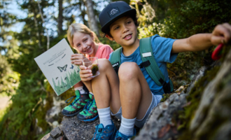 Zwei Kinder sitzen auf einem Stein in der Natur. Eines der Kinder hält ein Buch, während das andere ein Glas mit etwas in der Hand hat.