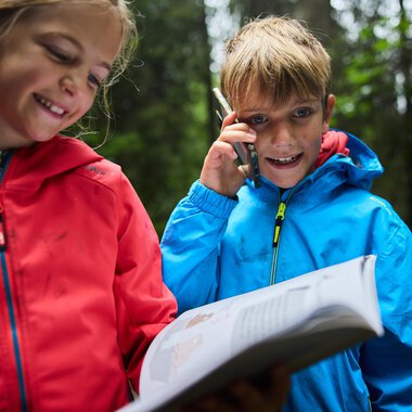Two children are standing in the forest. The girl is smiling while the boy is talking on the phone and holding a book.