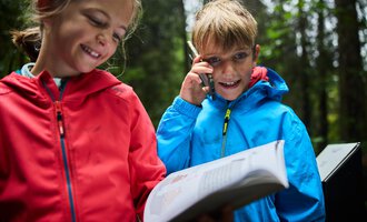 Two children are standing in the forest. The girl is smiling while the boy is talking on the phone and holding a book.
