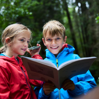 Two children are standing in the forest reading a book. They are wearing colorful rain jackets and laughing.