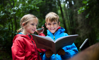 Two children are standing in the forest reading a book. They are wearing colorful rain jackets and laughing.