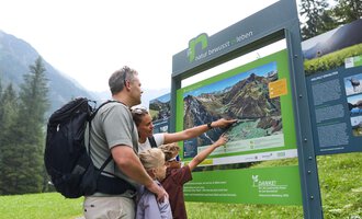 Eine Familie steht vor einem Informationsschild in der Natur. Sie zeigt auf die Karte und bespricht die Umgebung. | © Kleinwalsertal Tourismus | Oliver Farys