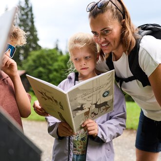 Eine Mutter liest mit ihren Kindern in der Natur. Die Kinder schauen neugierig in ein Buch. | © Kleinwalsertal Tourismus | Oliver Farys