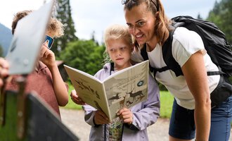 Eine Mutter liest mit ihren Kindern in der Natur. Die Kinder schauen neugierig in ein Buch. | © Kleinwalsertal Tourismus | Oliver Farys