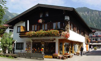 Ein traditionelles Haus in den Bergen, umgeben von Grün. Der Laden im Erdgeschoss bietet handgefertigte Holzschnitzereien an und ist mit Blumen geschmückt. | © Nebelhorn Holzschnitzerei e.K.