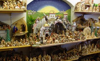 Eine detaillierte Krippe mit vielen Figuren und biblischen Szenen ist auf Regalen angeordnet. Im Hintergrund ist eine malerische Landschaft mit einem Himmel zu sehen. | © Nebelhorn Holzschnitzerei e.K.