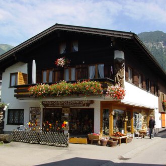 A traditional house in the mountains, surrounded by greenery. The shop on the ground floor offers handcrafted wooden carvings and is adorned with flowers. | © Nebelhorn Holzschnitzerei e.K.