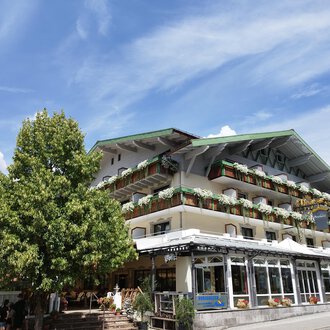 Ein charmantes Hotel mit Balkonen und bunten Pflanzen. Im Vordergrund steht ein großer Baum unter einem klaren blauen Himmel. | © Kleinwalsertal Tourismus | N. Lughammer
