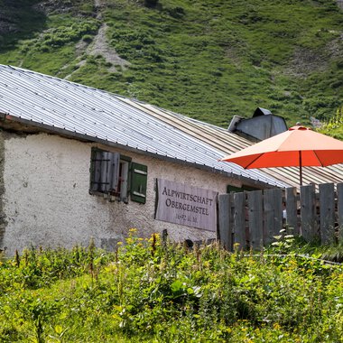 Eine charmante Almhütte mit einem roten Sonnenschirm im Grünen. Im Hintergrund sind sanfte Hügel und eine Berglandschaft zu sehen. | © Kleinwalsertal Tourismus | Andre Tappe