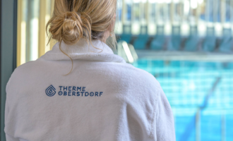 Eine Person in einem weißen Bademantel mit dem Logo "Therme Oberstdorf" steht vor einem Schwimmbecken. Es ist eine entspannende Atmosphäre sichtbar. | © Tourismus Oberstdorf