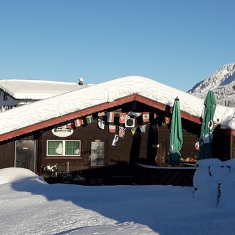 Ein gemütliches Holzhaus mit schneebedecktem Dach in einer winterlichen Landschaft. Im Hintergrund sind verschneite Berge und ein blauer Himmel zu sehen. | © Panoramahütte