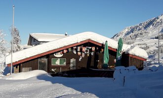 Ein gemütliches Holzhaus mit schneebedecktem Dach in einer winterlichen Landschaft. Im Hintergrund sind verschneite Berge und ein blauer Himmel zu sehen. | © Panoramahütte