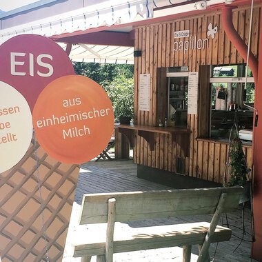 A cozy ice cream parlor with wooden paneling and a counter area. In the foreground, there is a large ice cream sign that indicates homemade ice cream. | © PAPILLON EISDIELE | Eis & Crêpe | Lisa Schüttler
