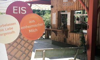 Eine gemütliche Eisdiele mit Holzverkleidung und einem Thekenbereich. Im Vordergrund steht ein großes Eisschild, das auf hausgemachtes Eis hinweist. | © PAPILLON EISDIELE | Eis & Crêpe | Lisa Schüttler