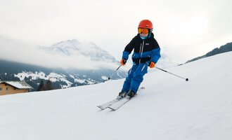 A boy is skiing on a snowy slope. In the background, mountains and clouds can be seen. | © Kleinwalsertal Tourismus | Oliver Farys