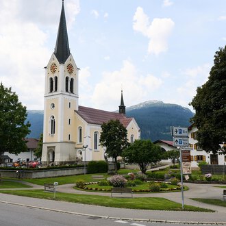 Eine ruhige Straße in einem charmanten Dorf mit einer hohen Kirche im Hintergrund. Umgeben von Bäumen und Bergen strahlt die Szene Ruhe aus. | © Kleinwalsertal Tourismus | Oliver Farys