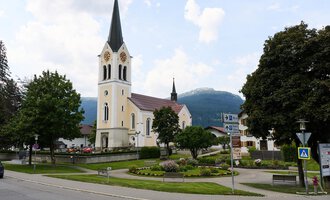 Eine ruhige Straße in einem charmanten Dorf mit einer hohen Kirche im Hintergrund. Umgeben von Bäumen und Bergen strahlt die Szene Ruhe aus. | © Kleinwalsertal Tourismus | Oliver Farys