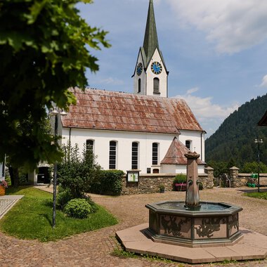 A picturesque village square with a fountain in the foreground and traditional houses. In the background, there is a church with a tall tower and a cloudy sky. | © Kleinwalsertal Tourismus | Oliver Farys