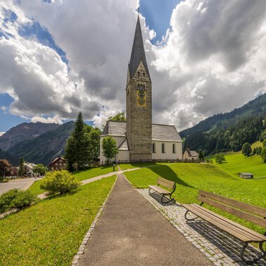 Eine malerische Kirche mit einem spitzen Turm steht in einer grünen Landschaft. Im Hintergrund sind sanfte Hügel und Wolken am Himmel zu sehen. | © Kleinwalsertal Tourismus | Steffen Berschin