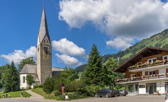 Eine malerische Straße mit einer Kirche und einem traditionellem Gebäude. Im Hintergrund sieht man grüne Hügel und einen blauen Himmel mit weißen Wolken. | © Kleinwalsertal Tourismus | Steffen Berschin