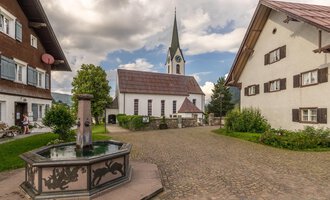 Ein malerisches Dorf mit traditionellen Häusern und einer Kirche im Hintergrund. In der Mitte befindet sich ein Brunnen, umgeben von grünem Gras und Bäumen. | © Kleinwalsertal Tourismus | Steffen Berschin