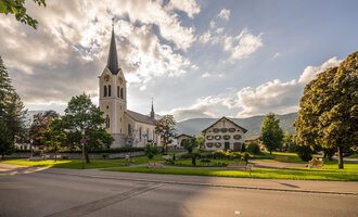 Eine malerische Kirche mit einem hohen Turm steht neben einem charmanten Gebäude. Der Himmel ist teilweise bewölkt und die Umgebung ist grün und einladend. | © Kleinwalsertal Tourismus eGen | Steffen Berschin