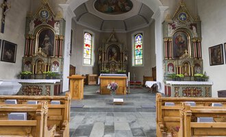 Ein schöner Kircheninneres mit Holzbankreihen und bunten Fenstern. Der Altar ist zentral platziert und von kunstvollen Verzierungen umgeben. | © Kleinwalsertal Tourismus | Oliver Farys