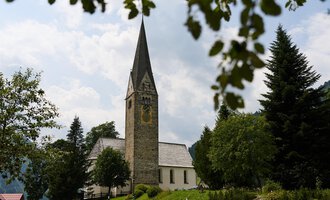 Eine malerische Straße mit einer Kirche und einem traditionellem Gebäude. Im Hintergrund sieht man grüne Hügel und einen blauen Himmel mit weißen Wolken. | © Kleinwalsertal Tourismus | Oliver Farys