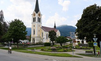 Eine malerische Kirche mit einem hohen Turm steht neben einem charmanten Gebäude. Der Himmel ist teilweise bewölkt und die Umgebung ist grün und einladend. | © Kleinwalsertal Tourismus | Oliver Farys