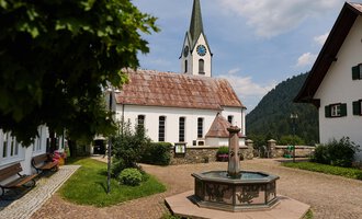 A picturesque village with traditional houses and a church in the background. In the middle there is a fountain, surrounded by green grass and trees. | © Kleinwalsertal Tourismus | Oliver Farys