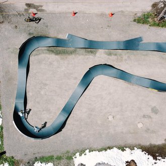 Eine Draufsicht auf eine geschwungene Rennbahn, die aus schwarzem Material besteht. Rund um die Bahn sind einige Menschen und Schneereste zu sehen. | © Christian Posch | Klaus Zopf